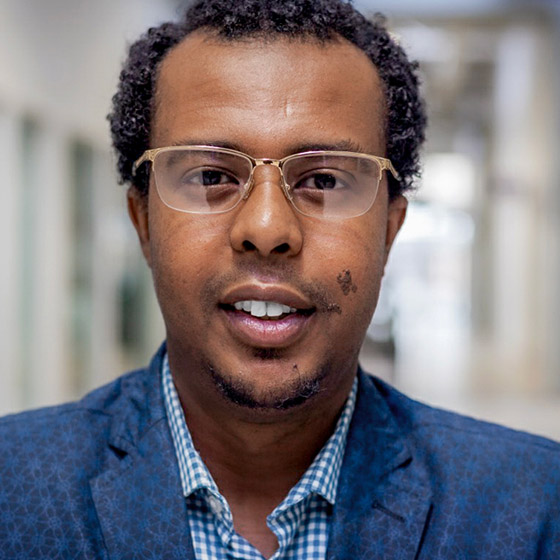 A man with short curly hair and glasses, wearing a blue patterned blazer and checkered shirt, stands indoors in a well-lit hallway, looking directly at the camera.