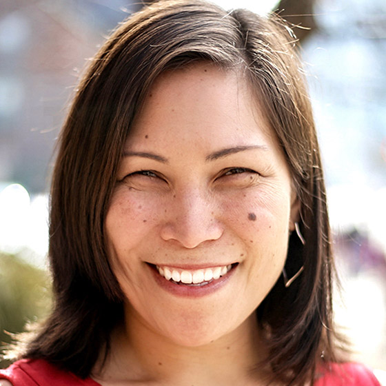 A woman with straight dark hair smiles warmly at the camera. She is outdoors with a blurred background, wearing a red top. Sunlight highlights her face, which has a few visible freckles and moles.