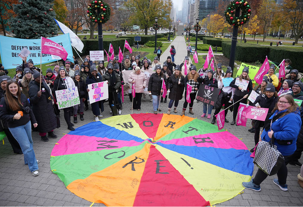 A large group of people hold colorful signs and flags, standing around a bright multicolored circle on the ground that reads WORTH MORE! in a city park with trees and buildings in the background.