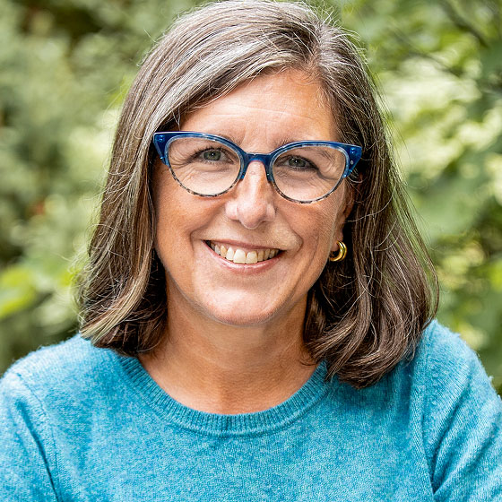 A middle-aged woman with shoulder-length gray hair, wearing blue glasses, gold hoop earrings, and a teal sweater, smiles at the camera with a green, blurred outdoor background.