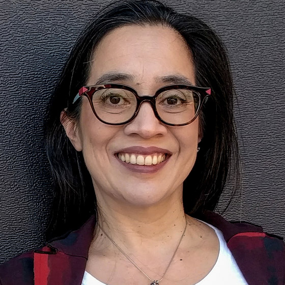 A woman with long dark hair and glasses smiles at the camera. She is wearing a white top, a red-and-black plaid shirt, and a necklace. The background is a dark, textured surface.