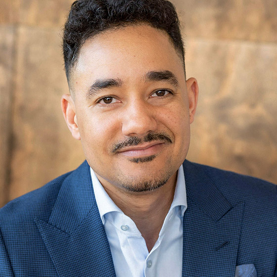A man with short curly hair and a trimmed goatee, wearing a blue suit jacket and light blue shirt, smiles slightly. The background is a blurred, warm brown tone.