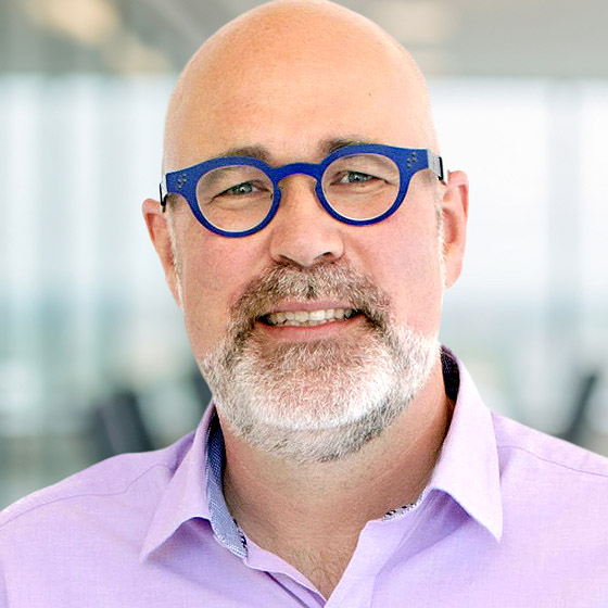 A smiling bald man with a beard and mustache, wearing blue round glasses and a light purple collared shirt, stands in a bright office environment with large windows in the background.