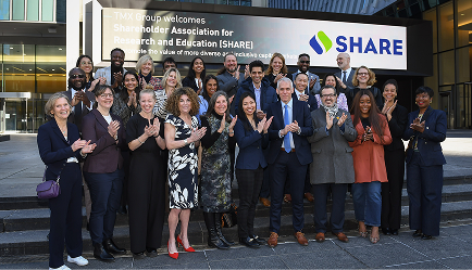 A large group of people smiling and posing for a photo in front of a building entrance with a sign that reads SHARE.